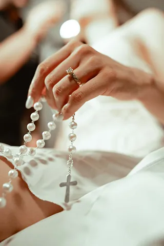Eine Braut hält mit der linken Hand, an der sich ein Ring befindet, eine Perlenkette mit Kreuz