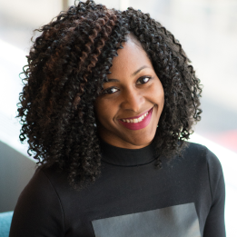 Lächende Frau mit lockigem schwarzen Haar und schwarzem Pullover vor hellem Hintergrund