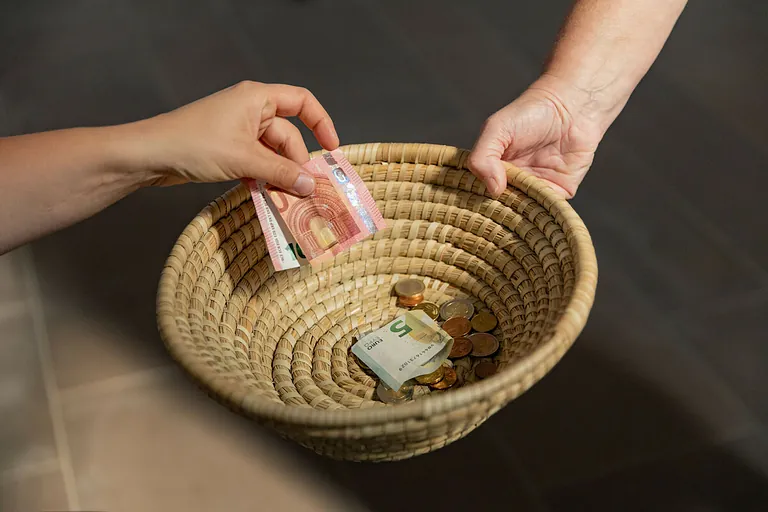 Eine Hand legt einen Geldschein in einen geflochtenen Korb; darin weitere Geldscheine und Münzen