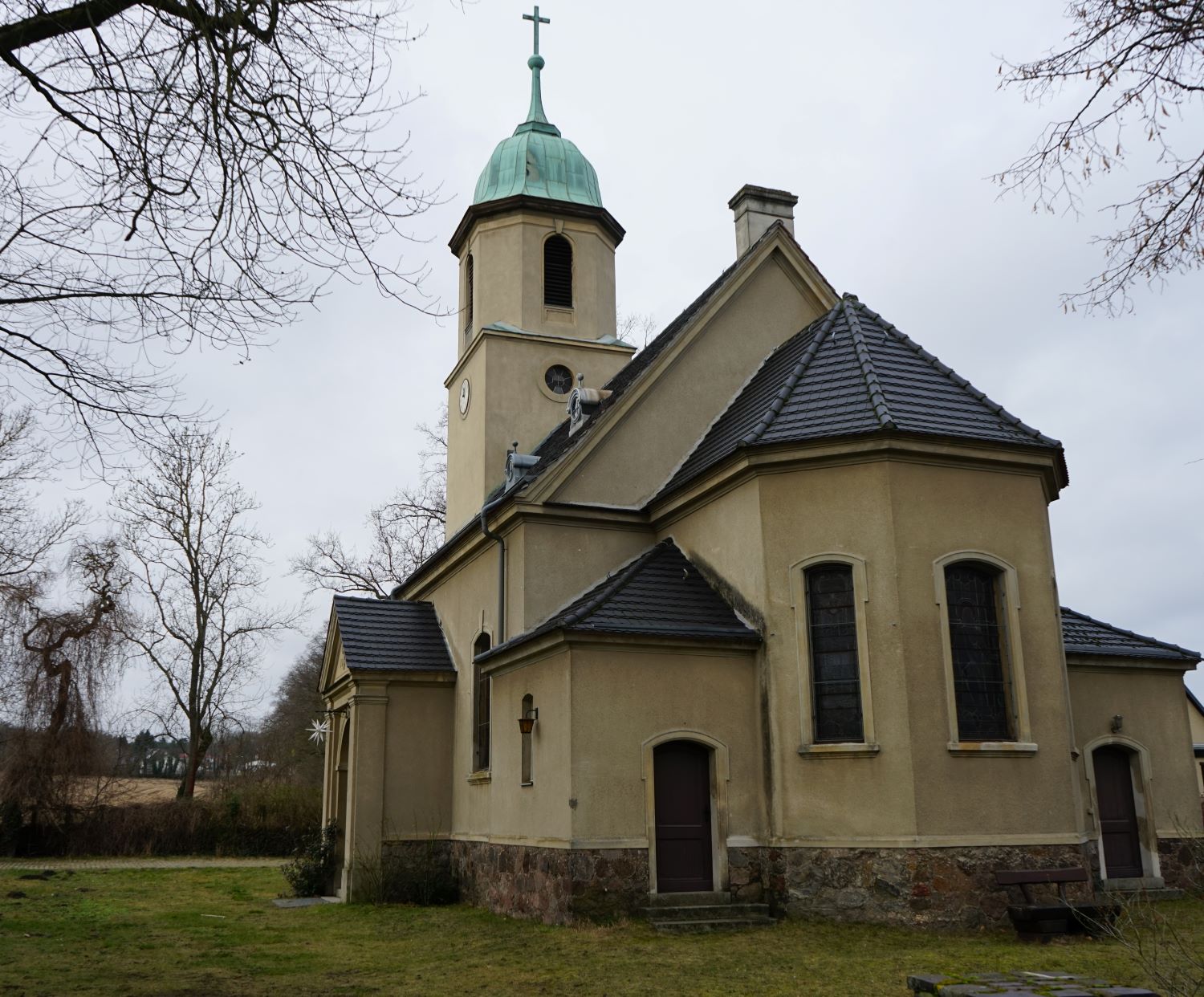 Kirchengebäude in Rosengarten mit grünem Turmdach und Kreuz, umgeben von Bäumen.