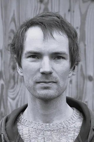 Junger Mann mit kurzem Haar in Pullover und Kapuzenjacke vor einer Holzwand