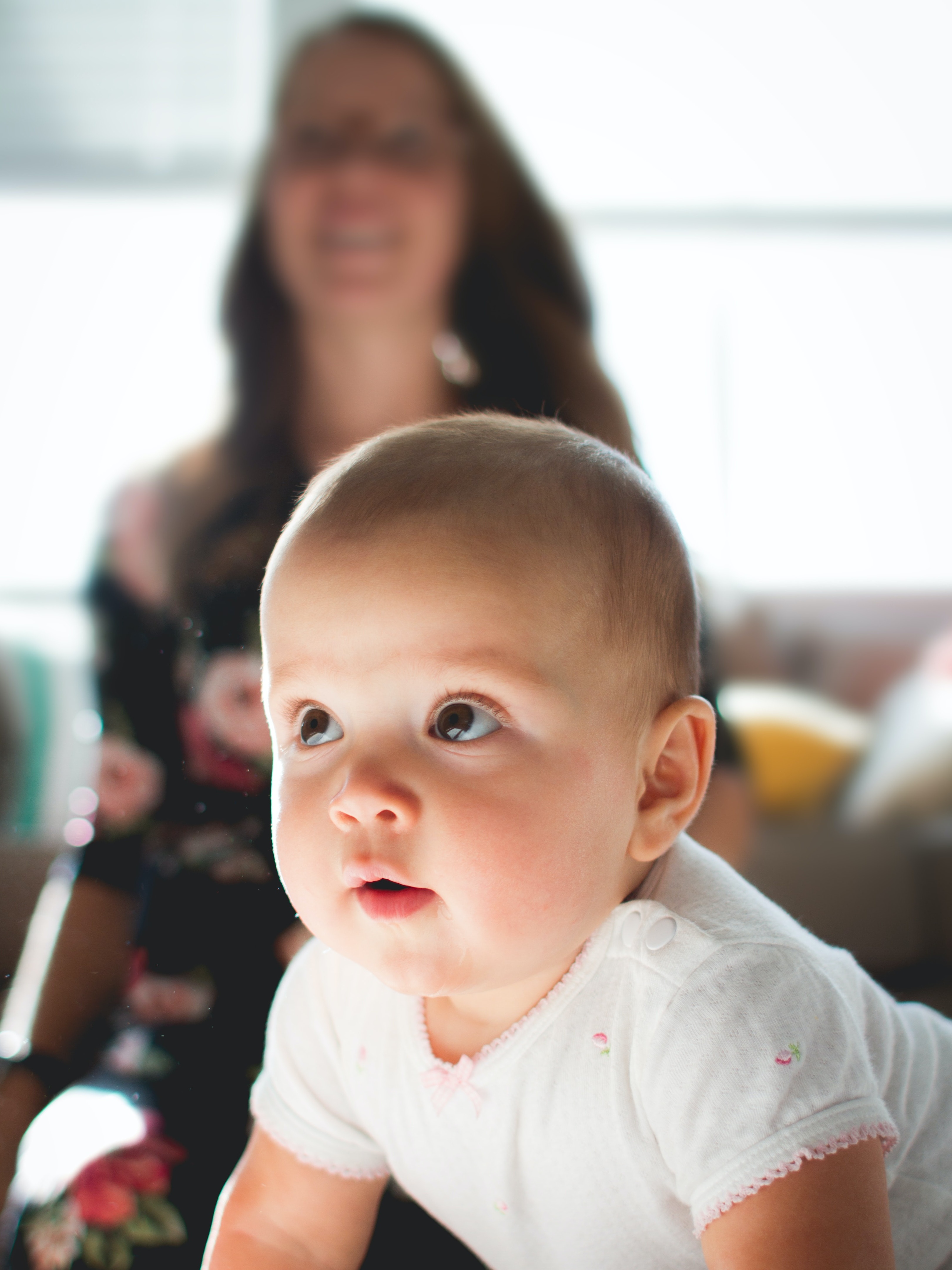 Nahaufnahme eines Babys mit weißem Oberteil. Im Hintergrund unscharf eine Frau mit dunklen Haaren.