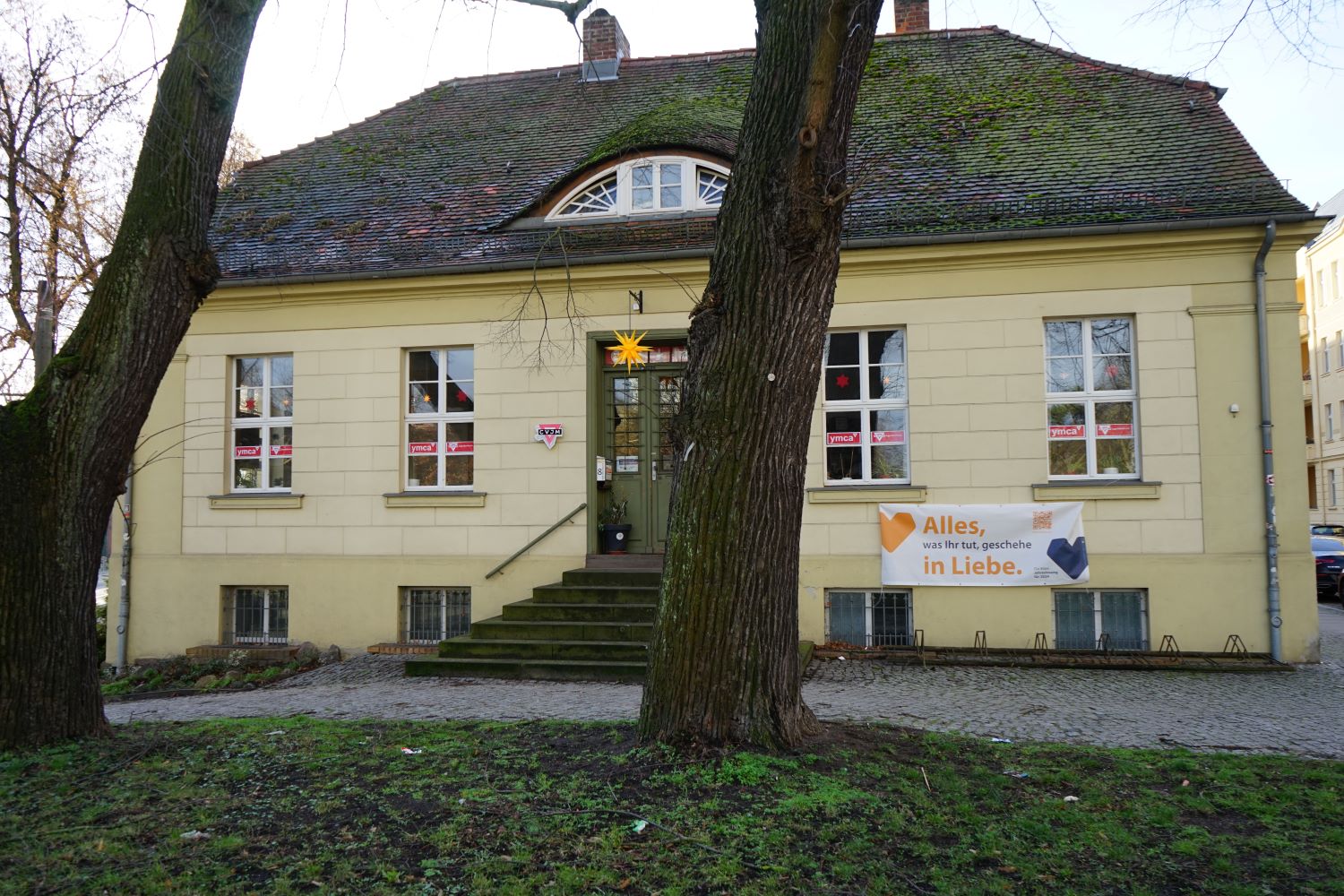 Gelbes Gebäude mit Treppe hinter zwei großen Bäumen. Sitz des CVJM in der Lindenstraße 8.