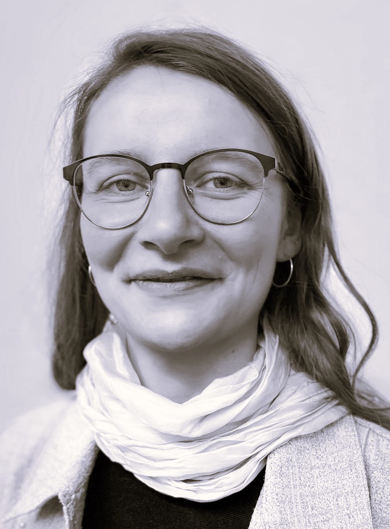 Junge Frau mit langen, gewellten Haaren und Brille in hellem Kostüm mit weißem Schal.